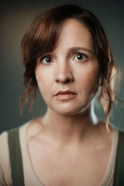 Lia in an olive pinafore and cream top, with a serious expression